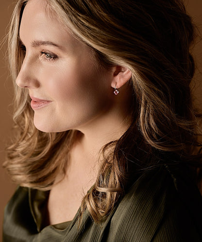 A woman in profile wearing a small pink gemstone dangle earring made from pure medical grade titanium with a golden ceramic coating, shown in a close-up portrait against a warm brown background.