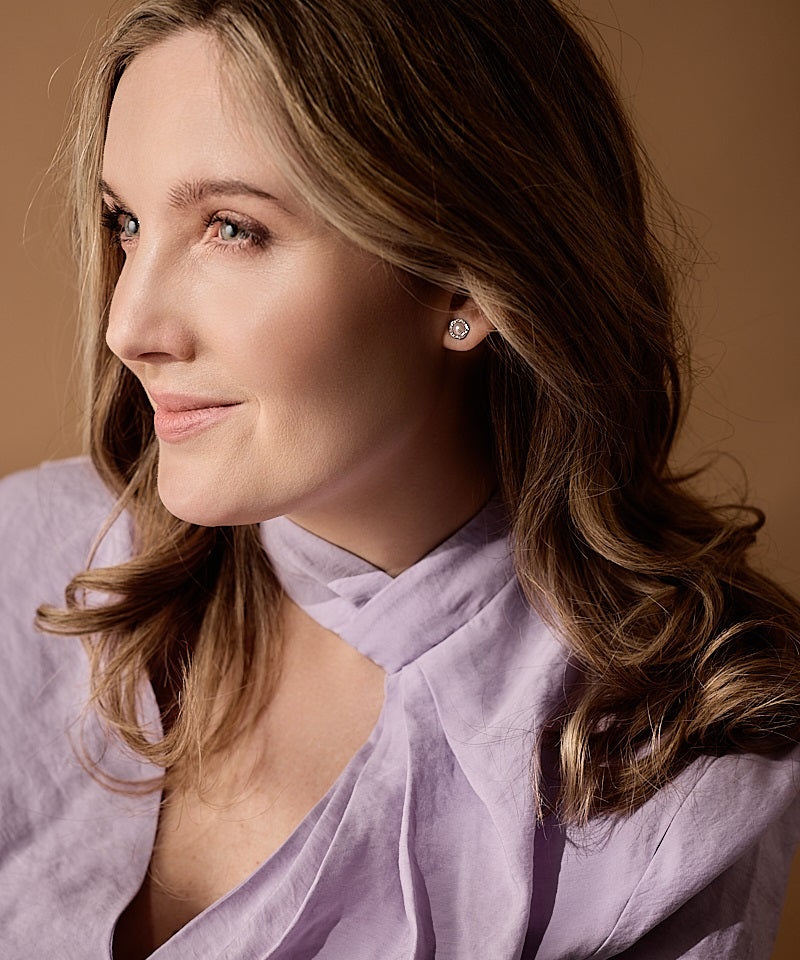 Woman wearing a light purple top with a soft brown background wearing a pair of Silver Titanium Brilliance 8mm Fancy Light Rose Pearl earrings surrounded by sparkling crystals.
